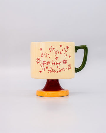 cream short ceramic mug with pink cursive 'in my growing season' with green handle and pink and yellow pedestal 