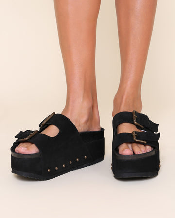 model wearing black suede platform slide sandals with oversized buckles and grommets