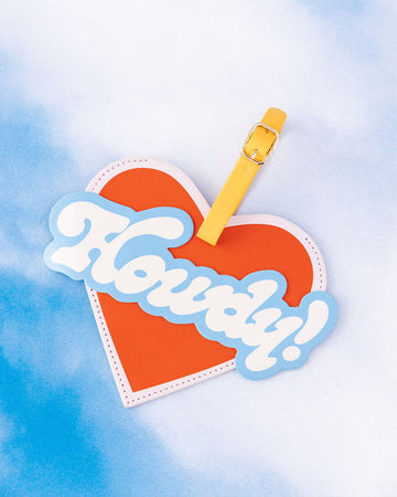 editorial image of red heart shaped luggage tag with blue and white 'Howdy!' in bubble letters